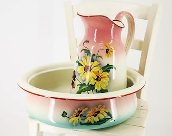 May include: A vintage enamelware pitcher and basin set with a floral design. The pitcher is pink with yellow flowers and green leaves. The basin is white with a pink and blue ombre effect and yellow flowers.