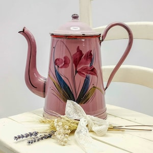 May include: A pink enamel teapot with a floral design. The teapot has a handle and a spout. It is sitting on a white chair with a white lace bow and dried flowers.