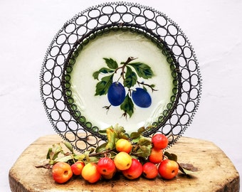May include: A vintage ceramic plate with a green and white design featuring two blue plums on a branch. The plate is surrounded by a black wire ring and sits on a wooden surface with a cluster of red and yellow apples.