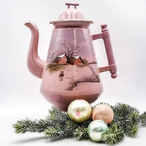 May include: A pink enamel teapot with a winter scene painted on it. The teapot has a lid with a knob and a handle. The scene includes houses, trees, and a person walking in the snow.