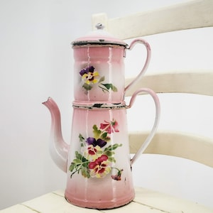 Könnte beinhalten: Eine rosafarbene Emaille-Kaffeekanne mit Blumenmuster. Die Kanne besteht aus zwei Teilen, einem oberen Teil zum Brühen und einem unteren Teil zum Aufbewahren des Kaffees. Die Kanne hat an jeder Seite einen Griff und einen Ausguss an der Seite.