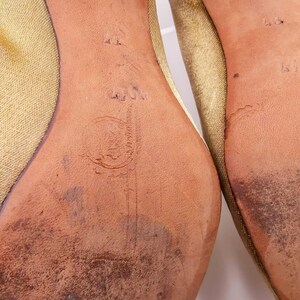 May include: Close-up of the bottom of a pair of gold satin shoes. The soles are leather and have the size 40 and the brand name "L.L.Jim" printed on them.