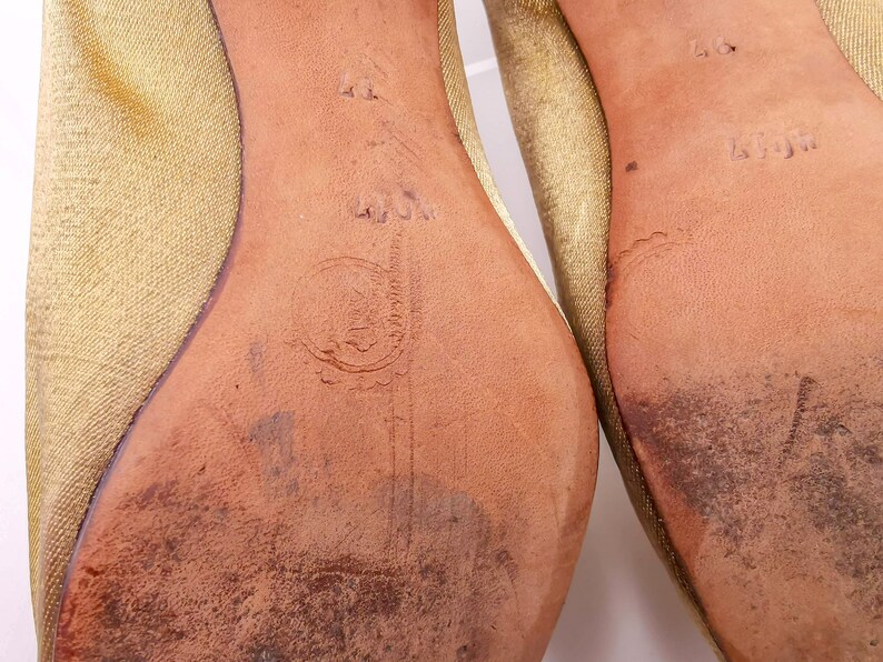 May include: Close-up of the bottom of a pair of gold satin shoes. The soles are leather and have the size 40 and the brand name "L.L.Jim" printed on them.