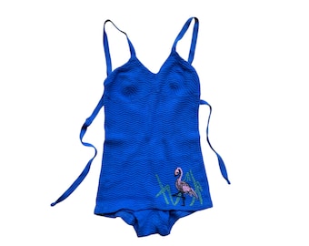 May include: A vintage blue swimsuit with a textured, chevron-like pattern. The one-piece suit features a V-neck and tie straps. A flamingo embroidery is on the lower front. The swimsuit is a vibrant shade of blue.
