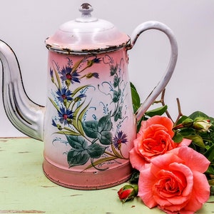 Könnte beinhalten: Eine vintage emaillierte Teekanne in Rosa und Weiß mit Blumenmustern, die auf einer grün-weiß gestrichenen Oberfläche mit rosafarbenen Rosen steht.
