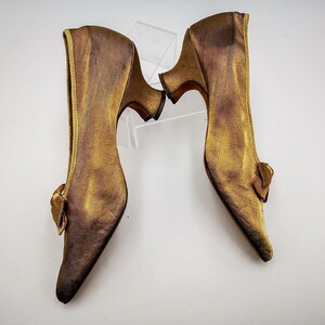 May include: A pair of gold satin heeled shoes with a bow detail on the back of each shoe.