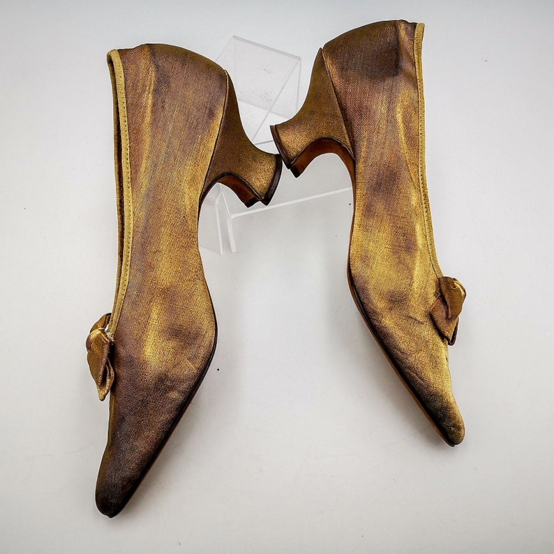 May include: A pair of gold satin heeled shoes with a bow detail on the back of each shoe.