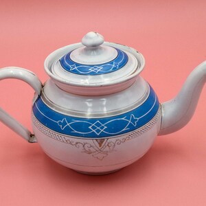 Può includere: Teiera in smalto bianco con un design decorativo blu e oro. La teiera ha un coperchio e un beccuccio.
