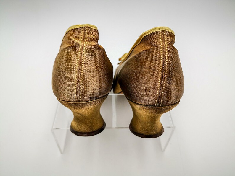 May include: A pair of gold satin heeled shoes with a pointed toe. The shoes are on a white background.