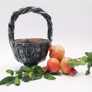 Puede incluir: Una cesta de metal negro con un diseño decorativo y un asa retorcida. Tres granadas rojas y naranjas están sentadas junto a la cesta con hojas verdes.