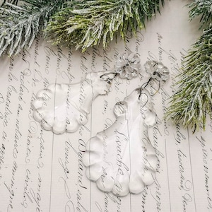 May include: Two clear glass crystal ornaments shaped like a teardrop with a scalloped edge. The ornaments are hanging from a silver wire.