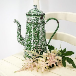May include: A green and white speckled enamel coffee pot with a lid and a handle. The pot is sitting on a white wooden chair with a bouquet of pink flowers.