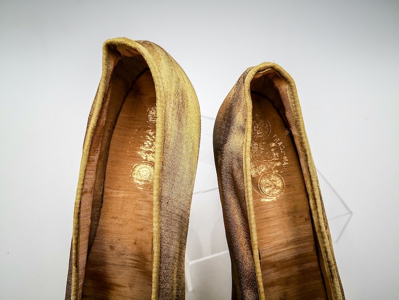 May include: A pair of gold satin slippers with a brown leather insole. The slippers have a pointed toe and a low heel.