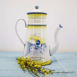 Könnte beinhalten: Eine weiße Emaille-Kaffeekanne mit blauem und gelbem Design. Die Kanne hat einen Ausguss und einen Griff. Das Design zeigt eine Bergszene mit einem Haus und Bäumen.