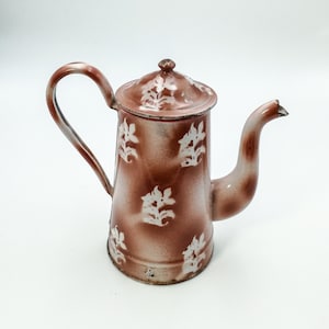 Könnte beinhalten: Eine braune Emailleteekanne mit einem weißen Blumenmuster. Die Teekanne hat einen Deckel und einen Ausguss.