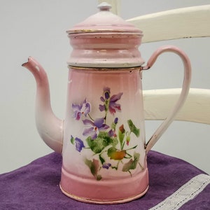 Könnte beinhalten: Eine Vintage-Kaffeekanne aus rosa Emaille mit Deckel und Griff. Die Kanne hat ein Blumendesign mit lila Blumen und grünen Blättern. Die Kanne steht auf einem lila Tuch.