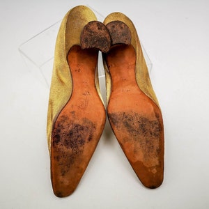 May include: A pair of gold satin heeled shoes with brown leather soles. The shoes are pointed toe and have a low heel.