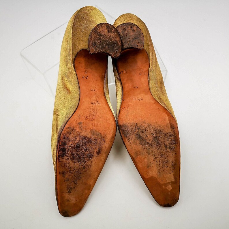 May include: A pair of gold satin heeled shoes with brown leather soles. The shoes are pointed toe and have a low heel.