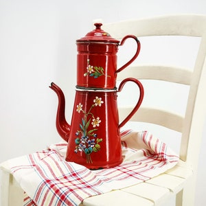 May include: A red enamel coffee pot with floral designs. The pot has a white lid and two handles. It is sitting on a white and red checkered cloth on a white chair.