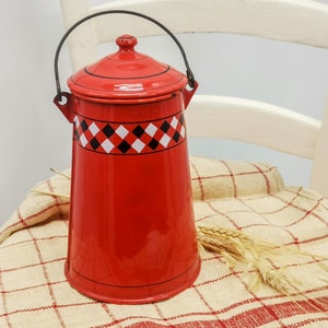 Könnte beinhalten: Eine rote Emaillekanne mit schwarzem Griff und Deckel. Die Kanne hat ein schwarz-weiß-rotes Karomuster am oberen Rand. Die Kanne steht auf einem karierten Tuch mit Weizenähren.