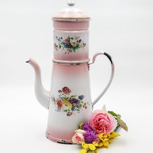 Könnte beinhalten: Eine antike Kaffeekanne aus rosa und weißem Emaille mit Blumendekor. Die Kanne hat einen herausnehmbaren Filter und einen Ausguss. Sie steht auf einer weißen Oberfläche mit rosa und gelben Blumen.