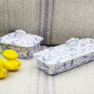Puede incluir: Dos cajas con tapa de cerámica blanca y azul con un diseño de escena pastoral. Una caja es rectangular, la otra es cuadrada. Las cajas están sobre una superficie de tono neutro con flores amarillas en primer plano.
