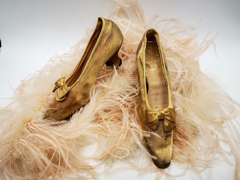 May include: A pair of gold satin shoes with a bow on the toe, resting on a bed of soft, light pink feathers.