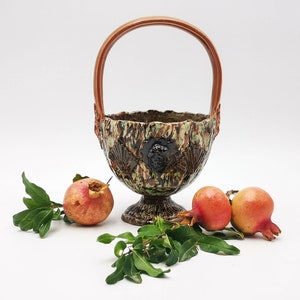 Puede incluir: Una cesta de cerámica con un asa marrón y un esmalte verde y marrón. La cesta está decorada con hojas en relieve y una cabeza de león. Hay tres granadas rojas junto a la cesta.