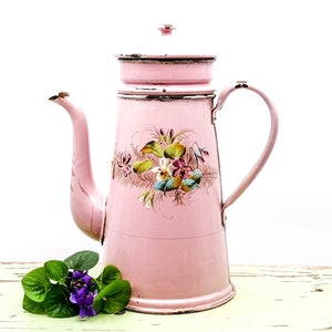 Könnte beinhalten: Eine antike Kaffeekanne aus rosa Emaille mit Blumenmuster. Die Kanne hat einen Deckel und einen Griff. Sie steht auf einer hellgrünen Holzoberfläche.