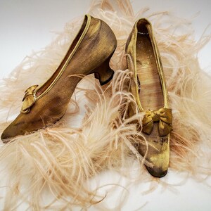 May include: A pair of gold satin shoes with a bow on the toe, sitting on a bed of soft, fluffy feathers.