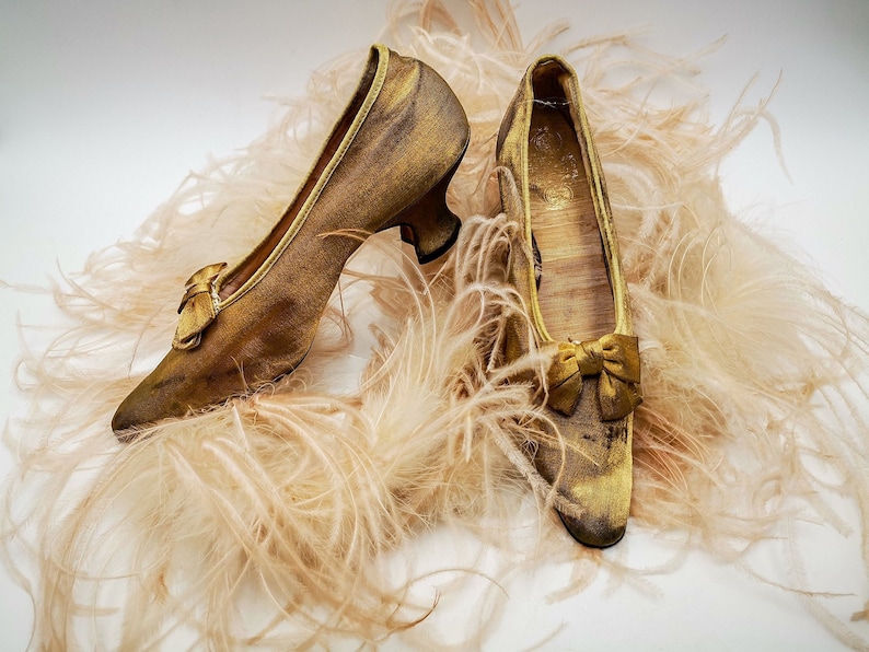 May include: A pair of gold satin shoes with a bow on the toe, sitting on a bed of soft, fluffy feathers.