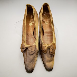 May include: A pair of gold satin shoes with a bow on the toe. The shoes are worn and have a vintage look.