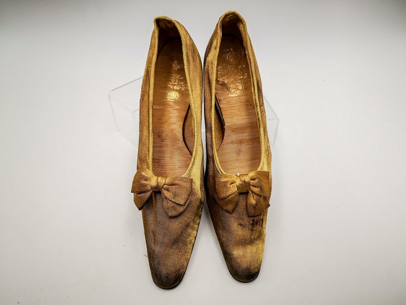 May include: A pair of gold satin shoes with a bow on the toe. The shoes are worn and have a vintage look.