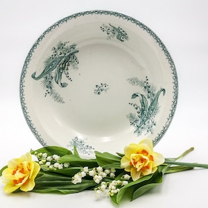 Puede incluir: Un cuenco de cerámica blanca con un diseño floral verde. El cuenco está decorado con un patrón de flores de lirio de los valles. El cuenco está sobre una superficie blanca con un ramo de narcisos amarillos y flores de lirio de los valles blancas.