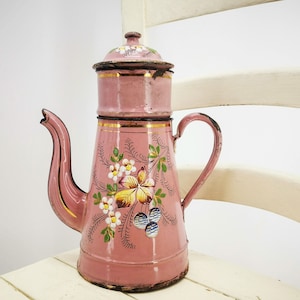 May include: A pink enamel coffee pot with a floral design. The pot has a gold trim and a lid with a floral design. The pot is sitting on a white surface.