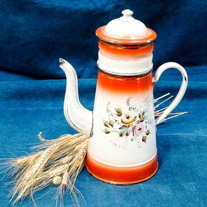 May include: A white and orange enamel coffee pot with a floral design. The pot has a lid and a handle. It is sitting on a blue surface with wheat stalks.