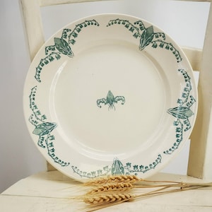 Peut inclure: Une assiette en céramique blanche avec un motif floral vert de muguet. L'assiette a un centre surélevé et un bord festonné.
