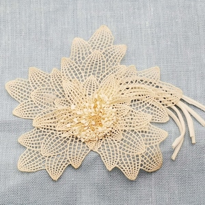 Può includere: Un delicato fiore di pizzo color crema con un centro testurizzato. Il fiore è fatto di fili intrecciati intricati e ha una sensazione delicata e ariosa.