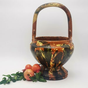 Puede incluir: Una cesta de cerámica marrón y negra con asa. La cesta está decorada con un patrón abstracto colorido. La cesta está sobre una superficie blanca con tres granadas rojas.