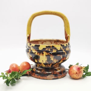 Puede incluir: Una cesta de cerámica amarilla y marrón con asa. La cesta está decorada con un patrón moteado. Tres granadas rojas están en primer plano.