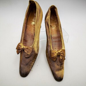 May include: A pair of gold satin shoes with a bow on the toe. The shoes have a pointed toe and a low heel.