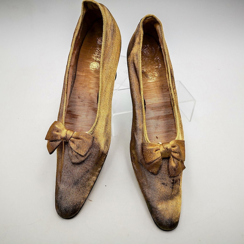 May include: A pair of gold satin shoes with a bow on the toe. The shoes have a pointed toe and a low heel.