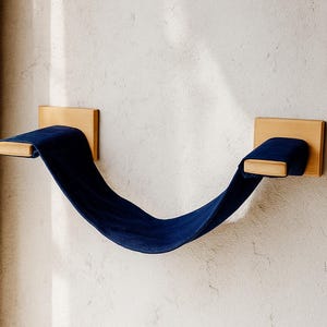 May include: A modern cat shelf with a curved, navy blue fabric suspended between two natural wood mounting blocks. The shelf is mounted on a textured, off-white wall, creating a minimalist design.