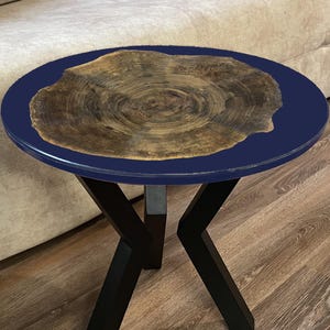 Puede incluir: Una mesa auxiliar redonda con una tapa azul oscuro y una incrustación de madera natural. La mesa tiene patas de metal negro y está colocada frente a un sofá beige. Una planta en maceta está a la izquierda de la mesa.
