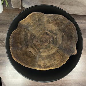 May include: A round black coffee table with a natural wood slab top. The wood slab is a light brown color with darker brown concentric rings. The table is a modern, rustic design.