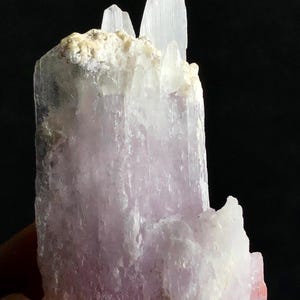 Peut inclure: Gros plan sur une formation cristalline violet clair avec des accents blancs et beiges. Le cristal a un aspect en couches avec plusieurs pointes. Le fond est noir.
