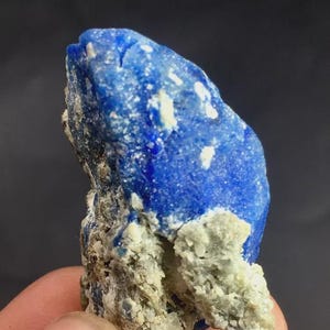 May include: A close-up of a piece of lapis lazuli. The stone is a deep blue color with white and gray inclusions. The rough texture of the stone is visible, with a mix of smooth and jagged surfaces. The stone is held in a person's hand.