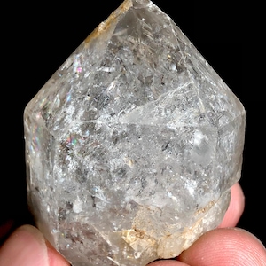 Puede incluir: Un cristal de cuarzo transparente con una punta y un cuerpo facetado. El cristal se sostiene en la mano de una persona sobre un fondo negro. El cuarzo tiene fracturas internas y algunas áreas de iridiscencia.