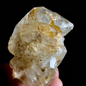Puede incluir: Un grupo de cristales de citrino natural. El cristal tiene un color amarillo pálido con áreas de tonos ámbar y marrón. La forma rugosa e irregular del cristal es visible sobre un fondo negro.