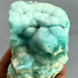 May include: A close-up of a mineral specimen with a layered, textured appearance. The stone features shades of blue-green and white, with a crystalline structure. The mineral is held in a hand, showcasing its natural form and color variations.